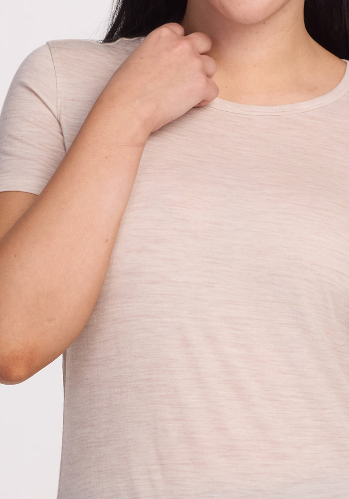 A person is seen from the neck to the waist wearing a Woolx Liza Crew Neck Tee in Cream Heather, made of lightweight fabric. Their left hand rests gently on their upper chest, and hair drapes partially over their shoulder against a plain white background.