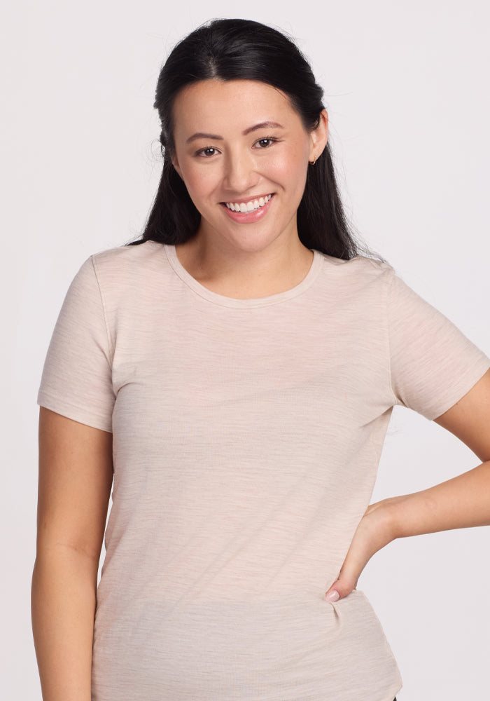 A person with long dark hair, wearing the Woolx Liza Crew Neck Tee in Cream Heather made from lightweight fabric, smiles against a light background with their right hand on their hip.