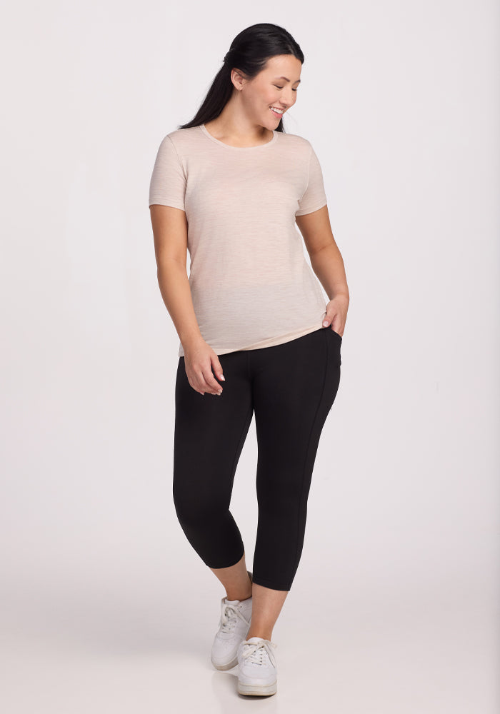 A person stands smiling, wearing a Woolx Liza Crew Neck Tee in Cream Heather and black leggings. They have long dark hair, white sneakers, and pose with one hand in their pocket against a plain backdrop.