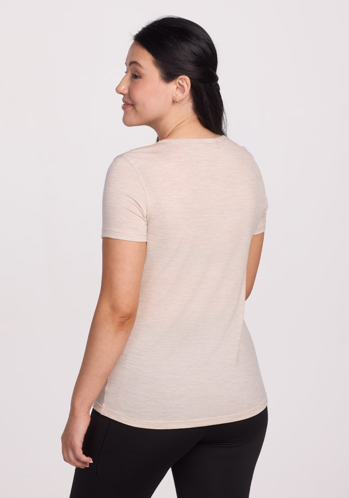 A person, smiling with their back to the camera, wears the Woolx Liza Crew Neck Tee in Cream Heather and black pants. The plain white background highlights the lightweight fabric's elegant versatility and performance benefits for any occasion.