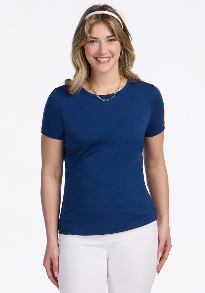 Woman smiling in a Woolx Liza Crew Neck Tee in Estate Blue, white pants, and gold necklace, on a white background.