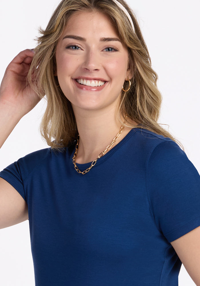 Smiling woman with wavy blonde hair in a Woolx Liza Crew Neck Tee in Estate Blue, gold hoops, and a chain necklace.