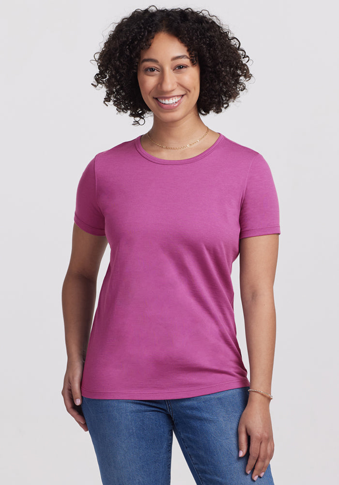Smiling woman in a Woolx Liza Crew Neck Tee in Purple Wine and blue jeans, standing against a white background.