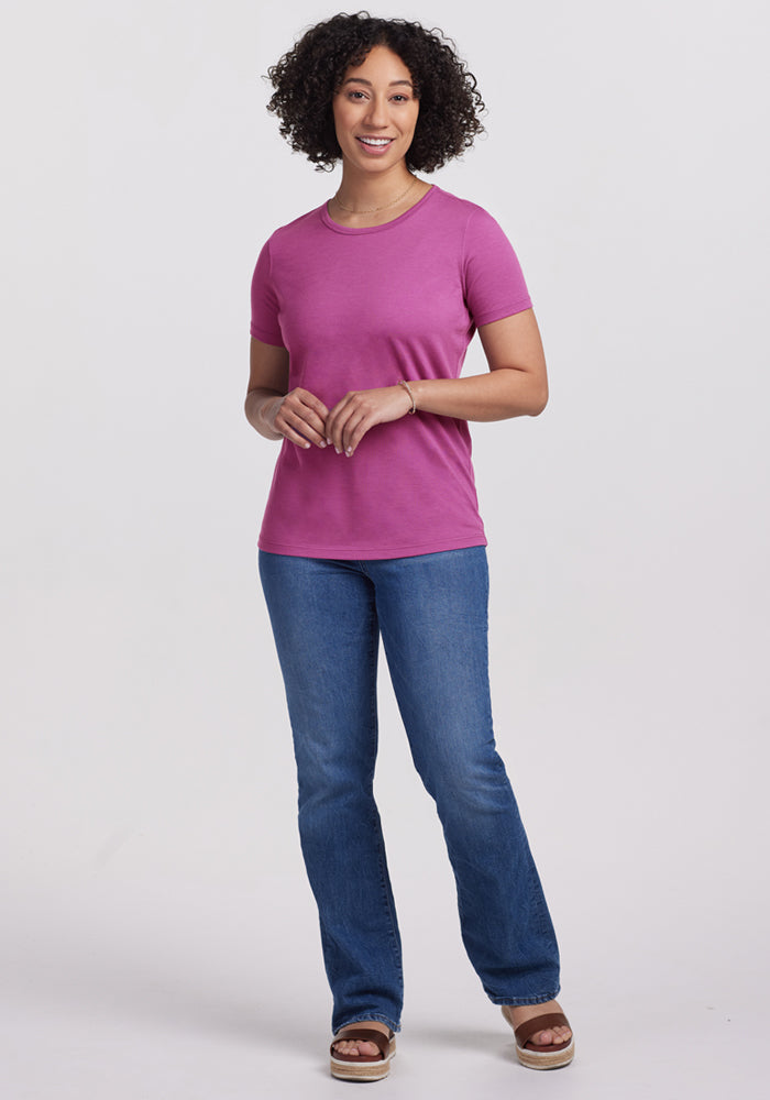 Smiling woman with curly hair wears Woolx Liza Crew Neck Tee in Purple Wine, blue jeans, and sandals, on white background.