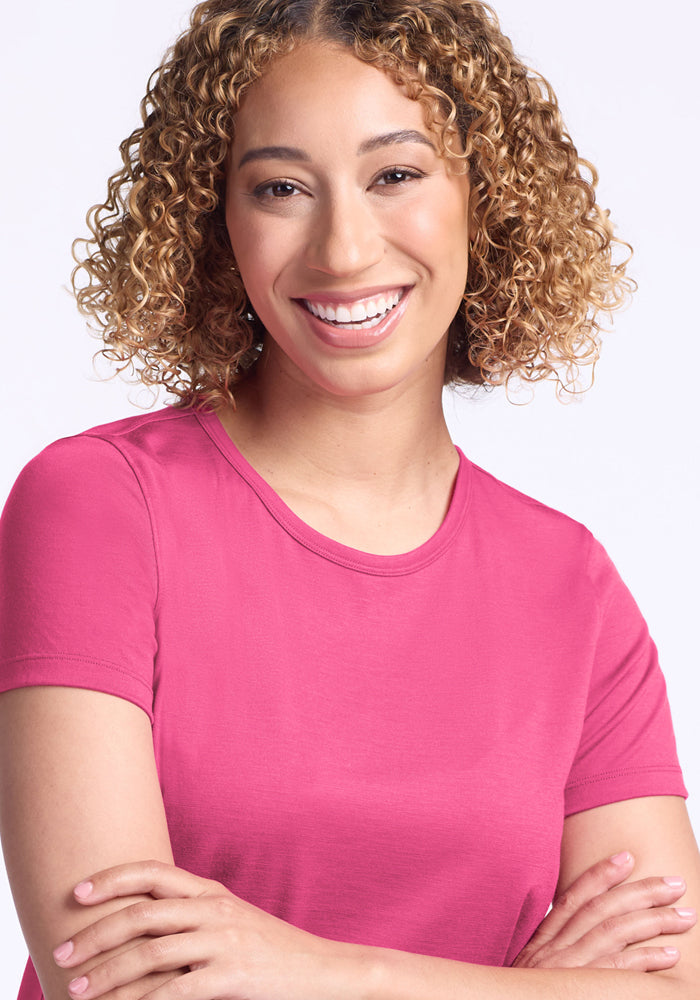 Smiling woman with curly hair wears the Woolx Liza Crew Neck Tee in Raspberry Fizz, arms crossed, against a light background.
