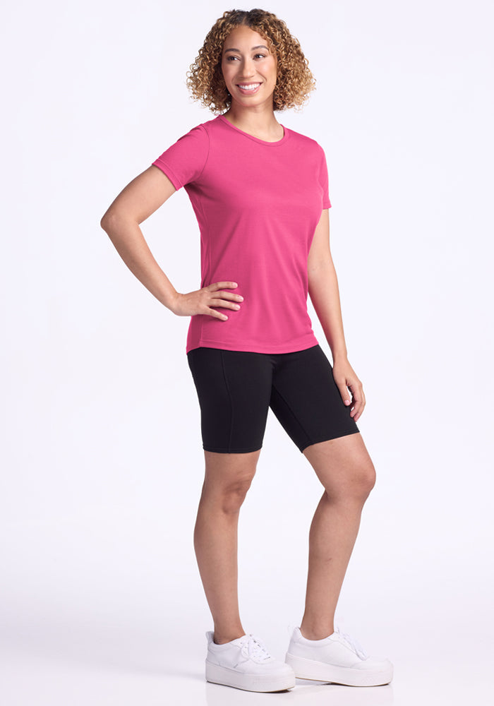 Woman in a Woolx Liza Crew Neck Tee in Raspberry Fizz and black shorts smiles with one hand on her hip.