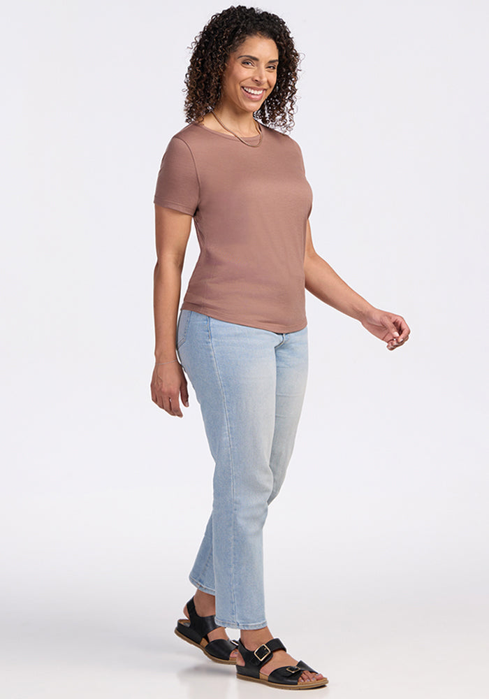 Woman with curly hair in a Woolx Liza Crew Neck Tee in Sienna Kiss, light blue jeans, and sandals smiles against a plain background.