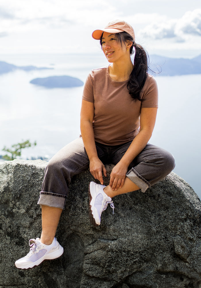 A woman in a Woolx Liza Crew Neck Tee in Sienna Kiss sits smiling on a rock with a lake and mountains behind her.