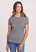 A person with shoulder-length brown hair, wearing the Woolx Liza Crew Neck Tee in Graphite Heather and blue jeans, stands against a plain white background, smiling gently. | Toni is 5’11”, wearing a size S