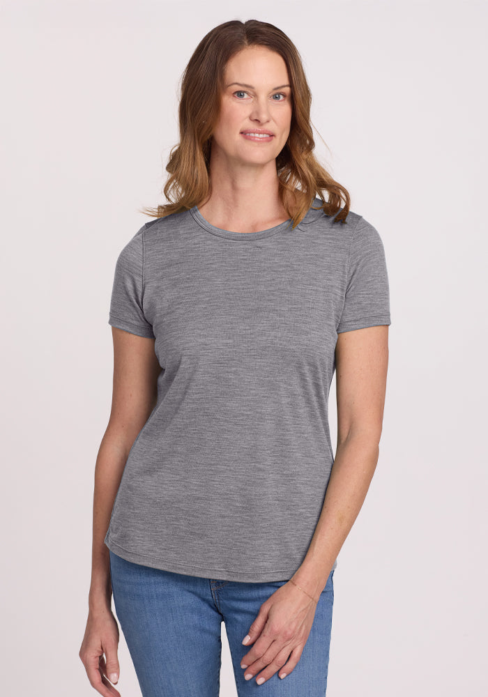 A person with shoulder-length brown hair, wearing the Woolx Liza Crew Neck Tee in Graphite Heather and blue jeans, stands against a plain white background, smiling gently. | Toni is 5’11”, wearing a size S
