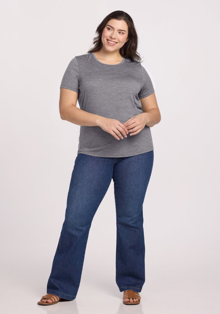 Smiling, a person stands in a Woolx Liza Crew Neck Tee in Graphite Heather, paired with blue jeans and brown sandals. They clasp their hands in front of them while posing against a plain white background.
