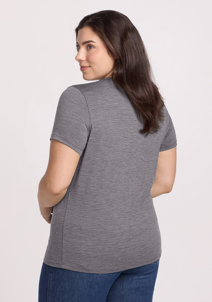 A person with long brown hair is turned slightly to the side, wearing a Woolx Liza Crew Neck Tee in Graphite Heather and blue jeans against a plain white background.