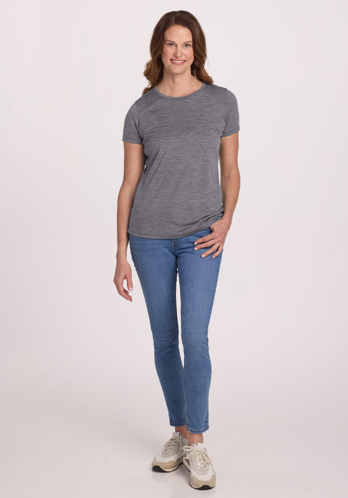 A person with long brown hair is wearing a Woolx Liza Crew Neck Tee in Graphite Heather, blue jeans, and white sneakers. They are smiling and standing against a plain white background.