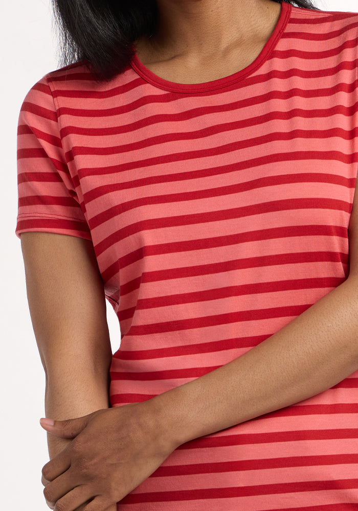A person with long dark hair wears the Woolx Liza Crew Neck Tee in Wildberry Stripe, featuring red and pink stripes and short sleeves. Their left hand rests on their right arm against a plain white background.
