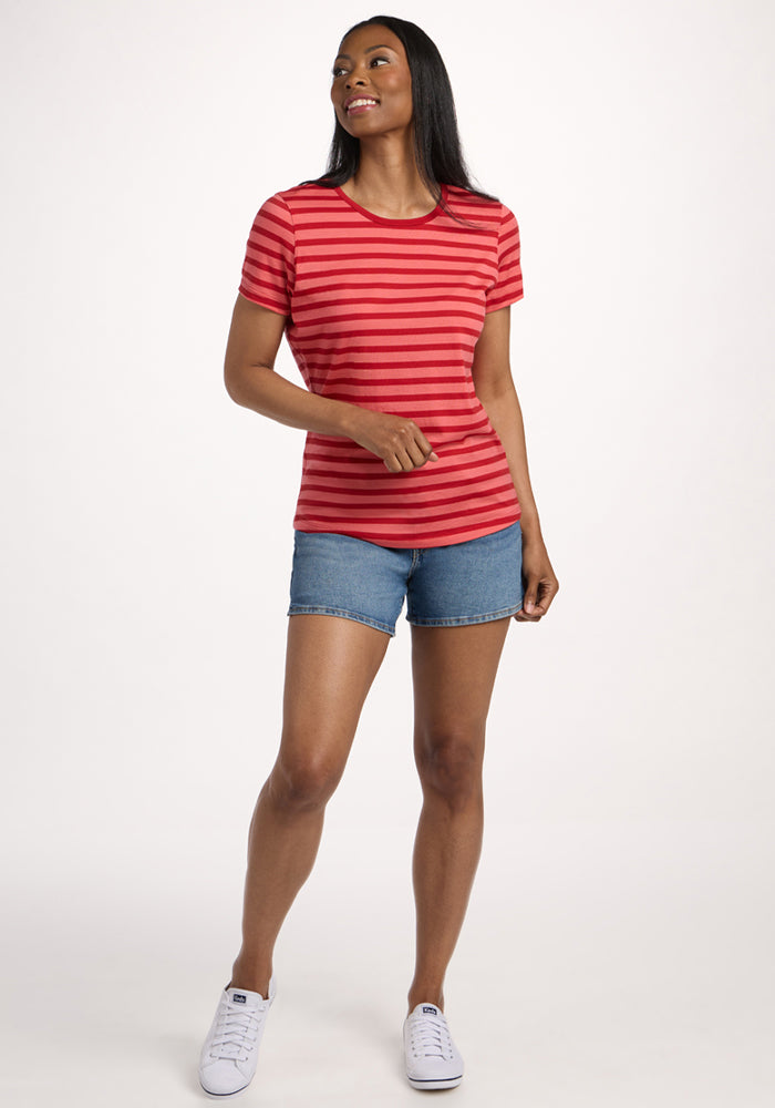 A woman smiles in the Woolx Liza Crew Neck Tee in Wildberry Stripe, paired with denim shorts and white sneakers. She has long straight hair and stands against a plain white background.