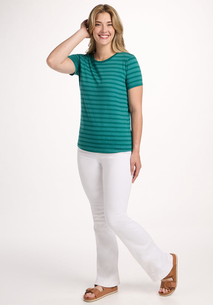 Smiling woman in Woolx Liza Crew Neck Tee - Woodland Stripe, white pants, and sandals stands against a white background.