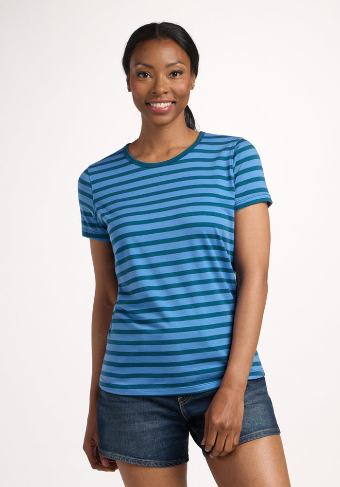 A woman stands against a plain background, smiling. She wears the Woolx Liza Crew Neck Tee - Coastal Stripe and denim shorts. Her hair is pulled back in a low ponytail. | Jade is 5’10”, wearing a size S