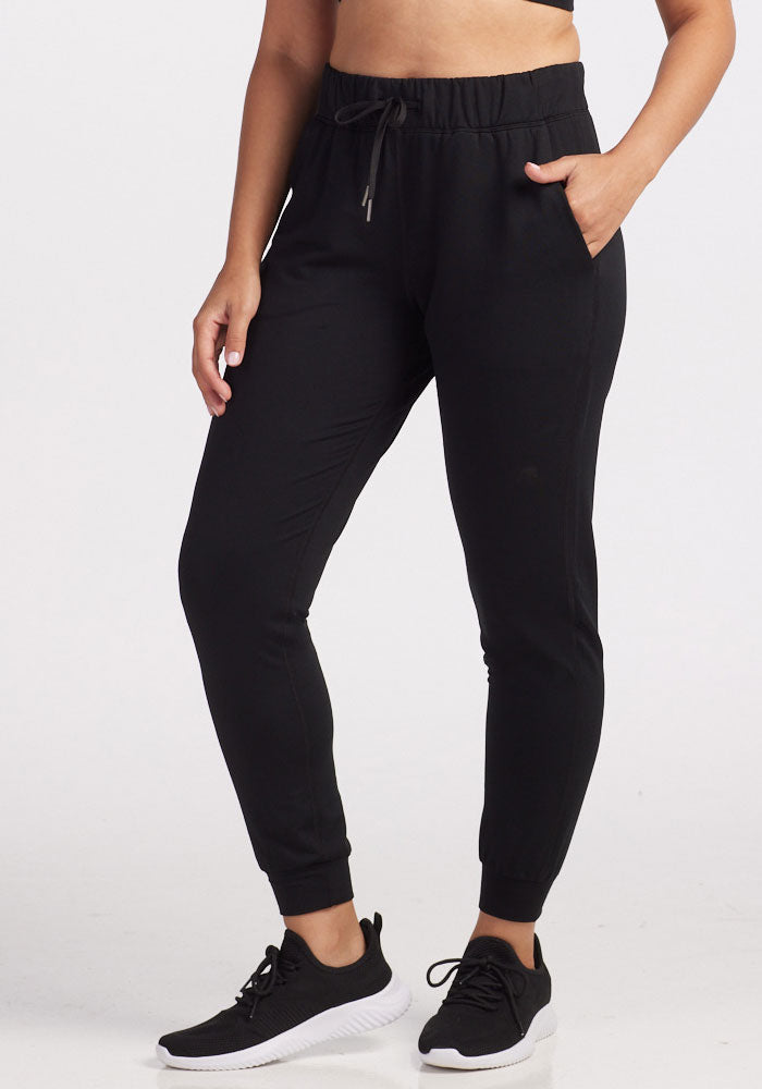 A person standing in travel-ready comfort, wearing Woolx's Lola Jogger in Black and black sneakers, casually has one hand in their pocket against a plain white background.