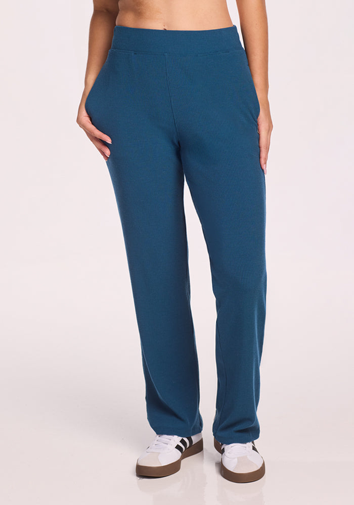 A person is wearing Woolx Luca Ribbed Pants in Moroccan Blue with white sneakers featuring brown soles. Their hands are in their pockets, shown waist down against a plain light background. | Ariana is 5'11", wearing a size S