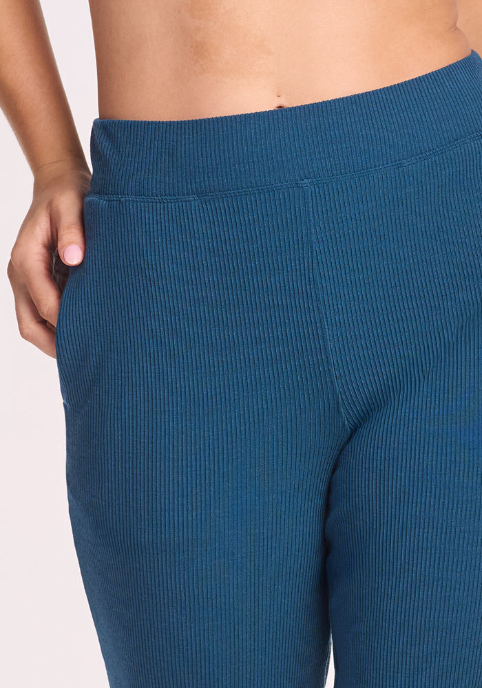 A close-up shows a person in Woolx Luca Ribbed Pants in Moroccan Blue with an elastic waistband, left hand in the pocket and their bare midriff visible.