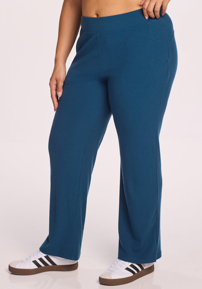 Someone wearing Woolx’s Luca Ribbed Pants in Moroccan Blue and white sneakers with black stripes stands with one hand on their hip against a plain, light background. | Juliana is 5'10", wearing a size XL