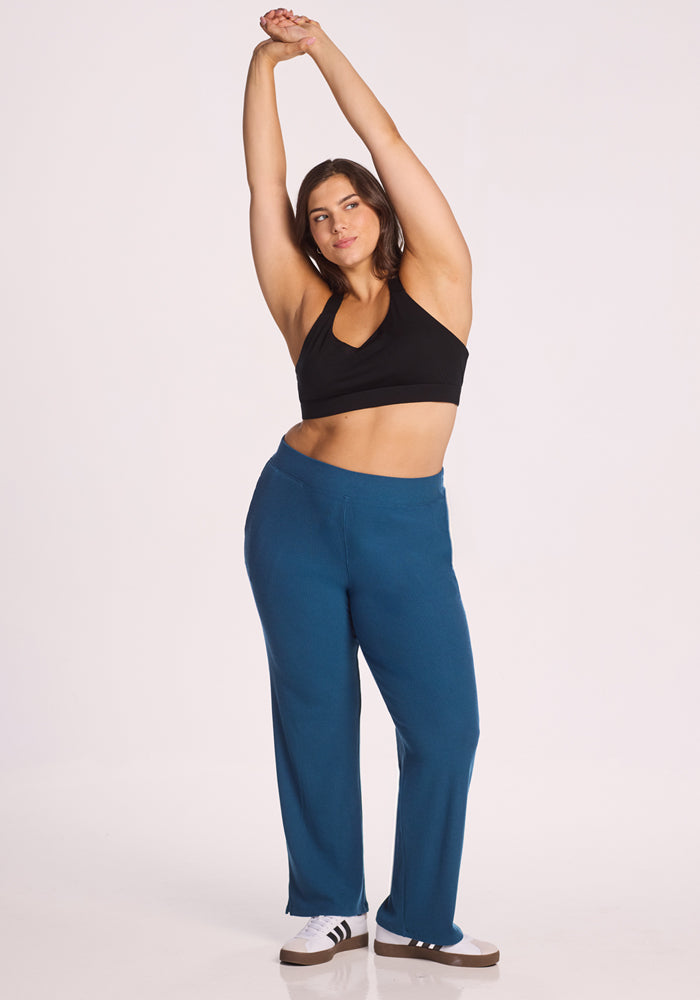 A woman wearing a black sports bra, Luca Ribbed Pants in Moroccan Blue by Woolx, and white sneakers with black stripes stands stretching with her arms raised against a plain white background.