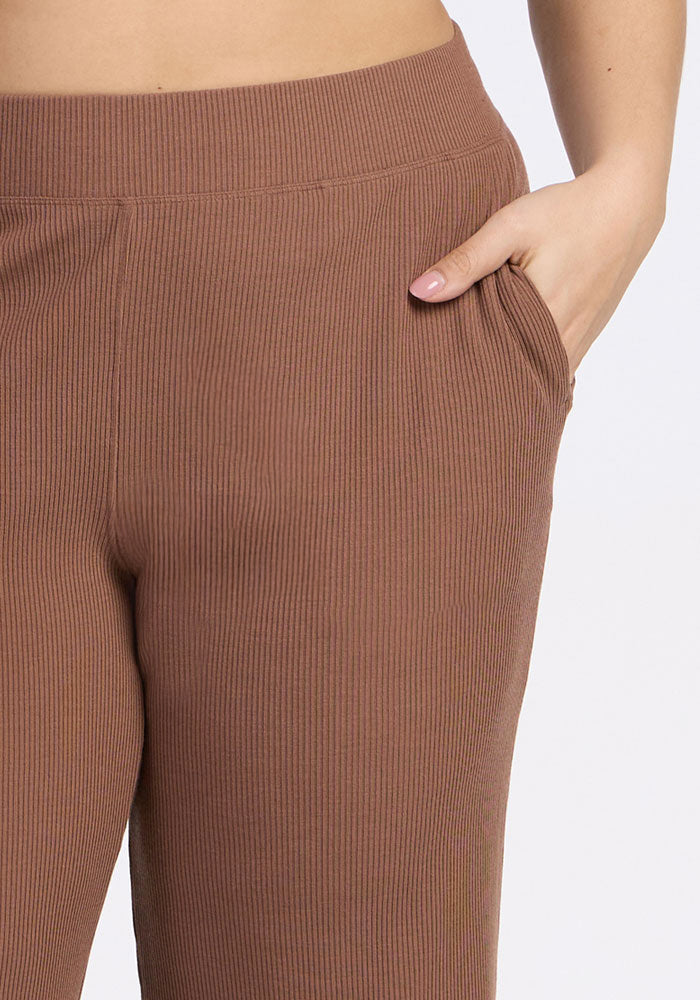 A person wearing Woolx's Luca Ribbed Pants in Sienna Kiss, high-waisted with wide legs and a hand in the right pocket, stands against a plain light background.