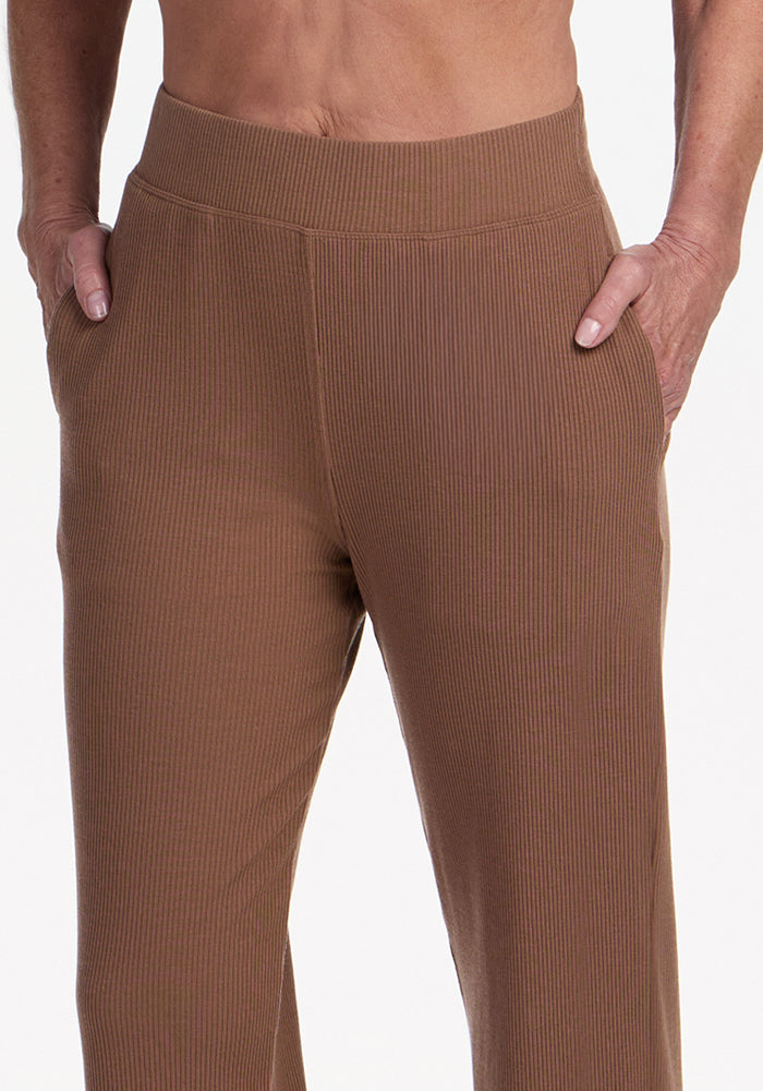 A person stands with hands in the pockets of the Woolx Luca Ribbed Pants in Sienna Kiss. The image is cropped below the chest, highlighting the cozy, textured high-waisted pants.