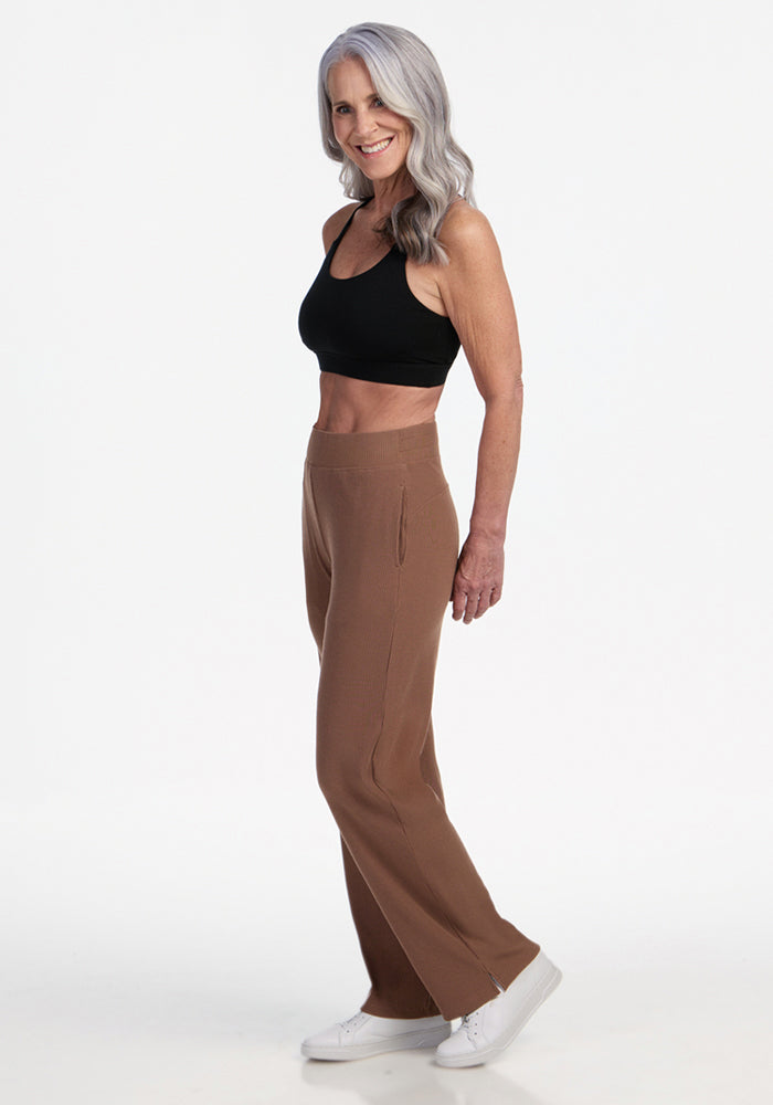 A smiling older woman with long gray hair stands sideways against a white background, wearing a black sports bra, Woolx Luca Ribbed Pants in Sienna Kiss, and white sneakers.