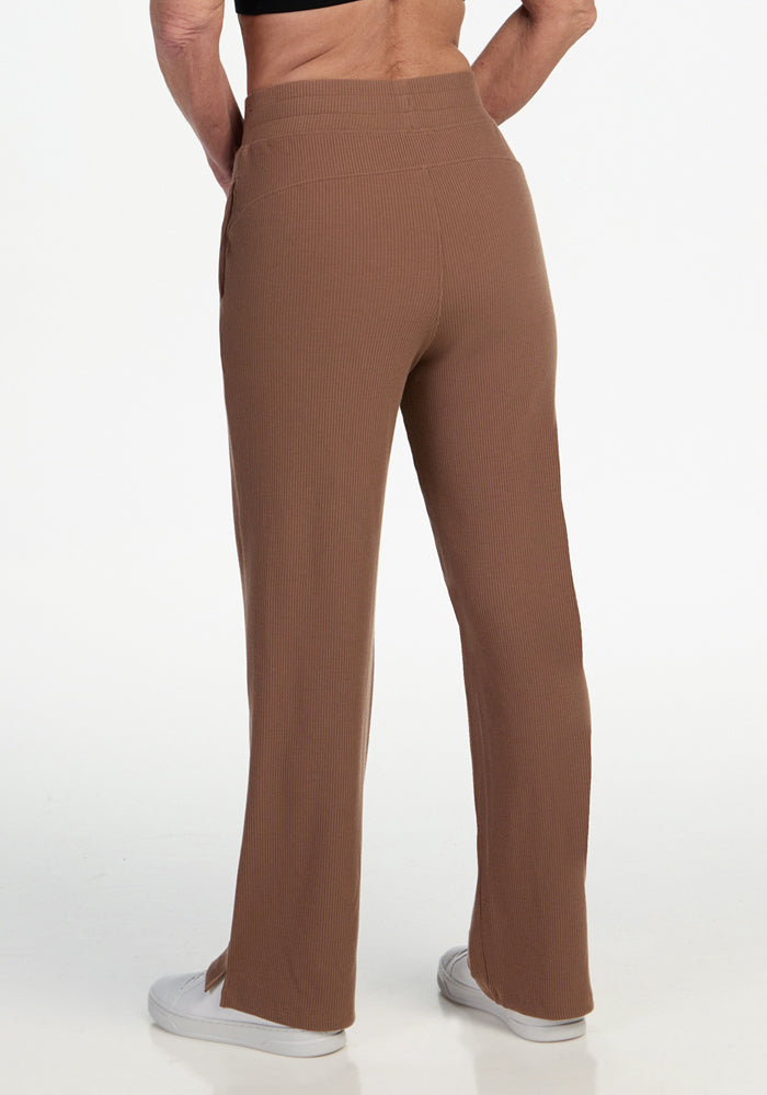 A person stands against a white background, back to the camera, wearing Woolx Luca Ribbed Pants in Sienna Kiss with wide legs and white sneakers, paired with a black top.