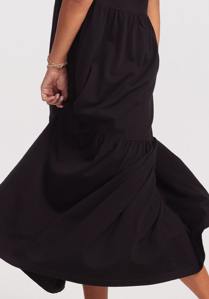A person twirls in a long, flowing Lucia Dress - Black by Woolx, causing the tiered ruffle fabric to billow. The emphasis is on the dress's lower half and a bracelet-adorned hand against a plain, neutral background.