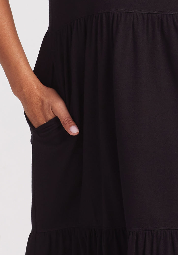 Close-up of someone in the Woolx Lucia Dress - Black, featuring a gathered skirt. Their right hand rests casually in the side pocket, highlighting the design and merino wool texture against a plain light background.
