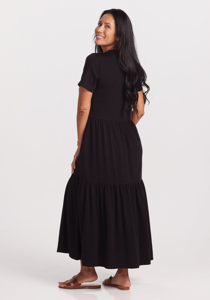 A woman with long black hair smiles over her shoulder in a Woolx Lucia Dress - Black, crafted from luxurious, temperature-regulating merino wool. She's wearing brown sandals against a plain white background.