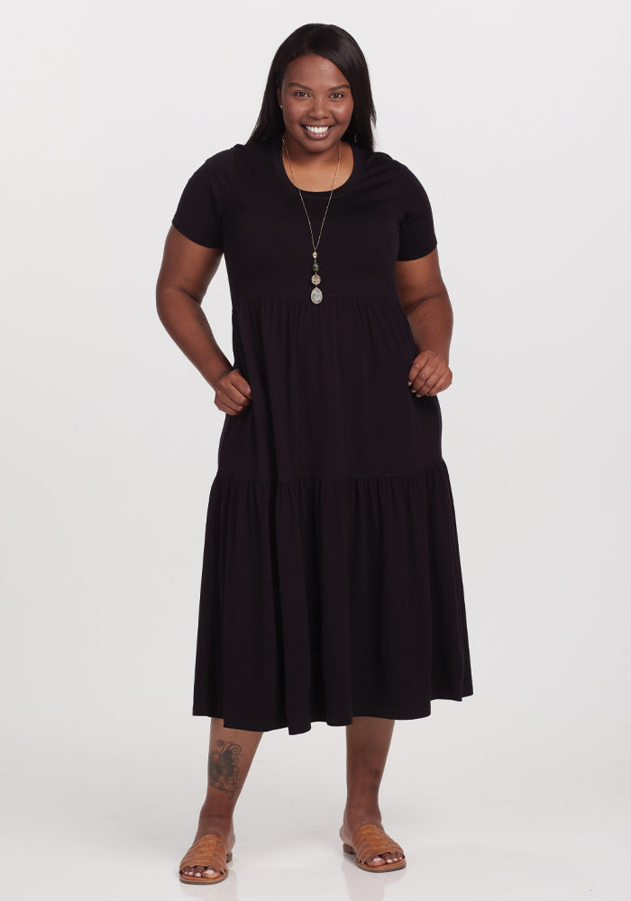 A woman smiles while posing in a black, short-sleeved Woolx Lucia Dress made of temperature-regulating merino wool. She accessorizes with a long necklace and sandals, her hands in the dress pockets, against a white backdrop. | Le'Quita is 5'11", wearing a size XL