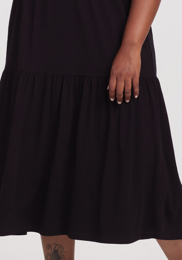 A person in a Woolx Lucia Dress - Black with a ruffled hem stands against a white background. Their right hand is by their side, and a tattoo shows above the left knee. The dress is made of temperature-regulating merino wool for comfort.