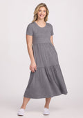 A person with long blonde hair wears a Woolx Lucia Dress in Graphite Heather, ideal for summer, paired with white sneakers. They stand against a plain white background, smiling with one hand in their pocket. | Ariana is 5’11”, wearing a size S