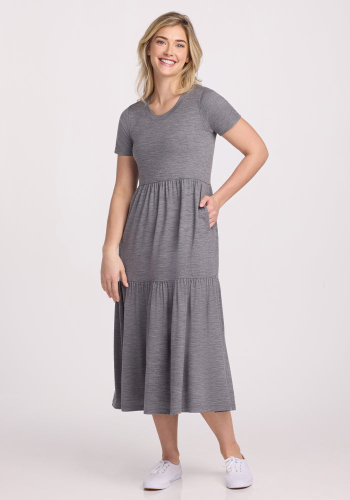 A person smiles in the Woolx Lucia Dress in Graphite Heather, made from breathable Merino wool. Paired with white sneakers against a plain background, one hand is in their pocket and the other at their side—perfectly dressed to beat the summer heat.