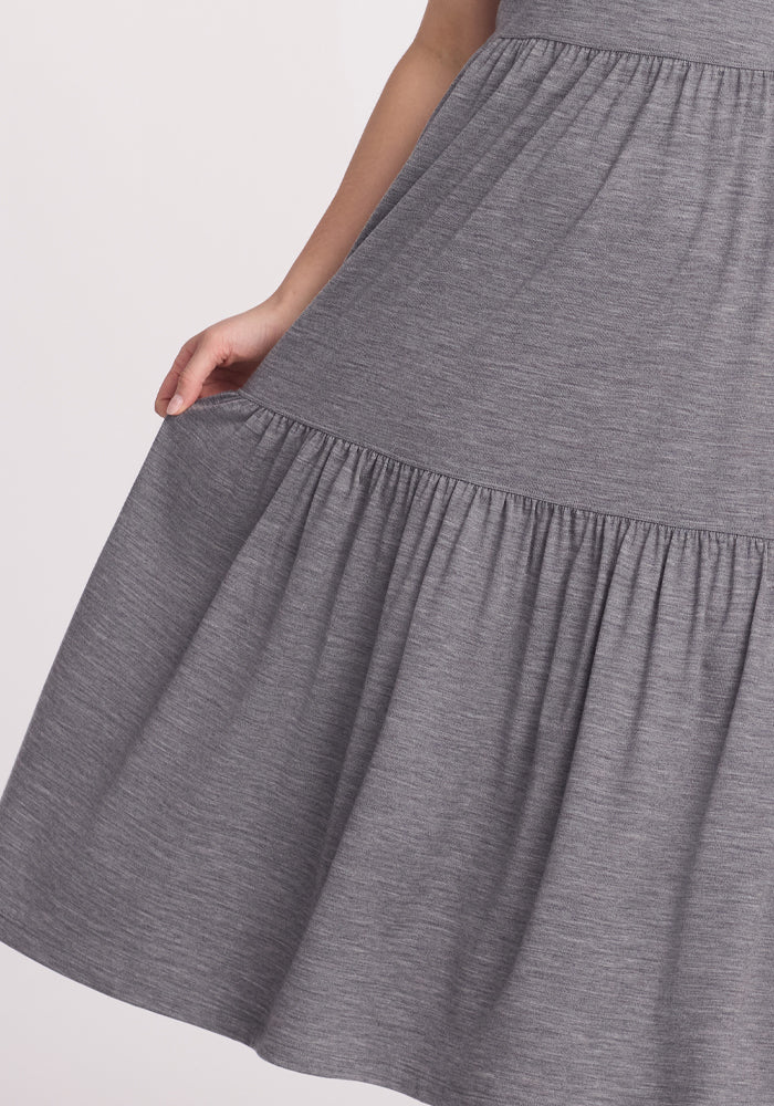 A person in a Woolx Lucia Dress - Graphite Heather, knee-length and made of merino wool with gathered fabric detail, lightly holds the side of the dress. The plain light-colored background adds cool elegance for summer days.