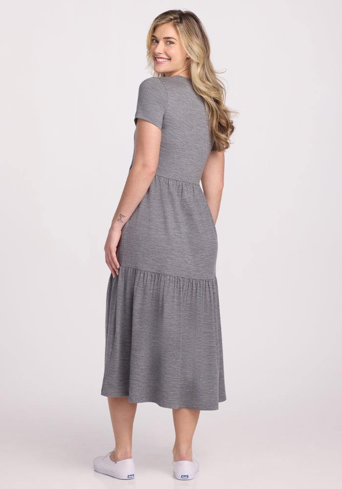 A woman with long blonde hair smiles while standing sideways, looking back. She wears a short-sleeved Lucia Dress in Graphite Heather by Woolx, crafted from merino wool, draping to her ankles and paired perfectly with white sneakers. The backdrop is plain white.