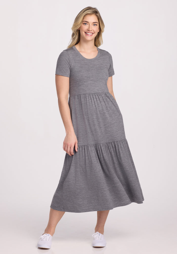 A person with long blonde hair wears a Woolx Lucia Dress in Graphite Heather, ideal for summer, paired with white sneakers. They stand against a plain white background, smiling with one hand in their pocket. | Ariana is 5’11”, wearing a size S