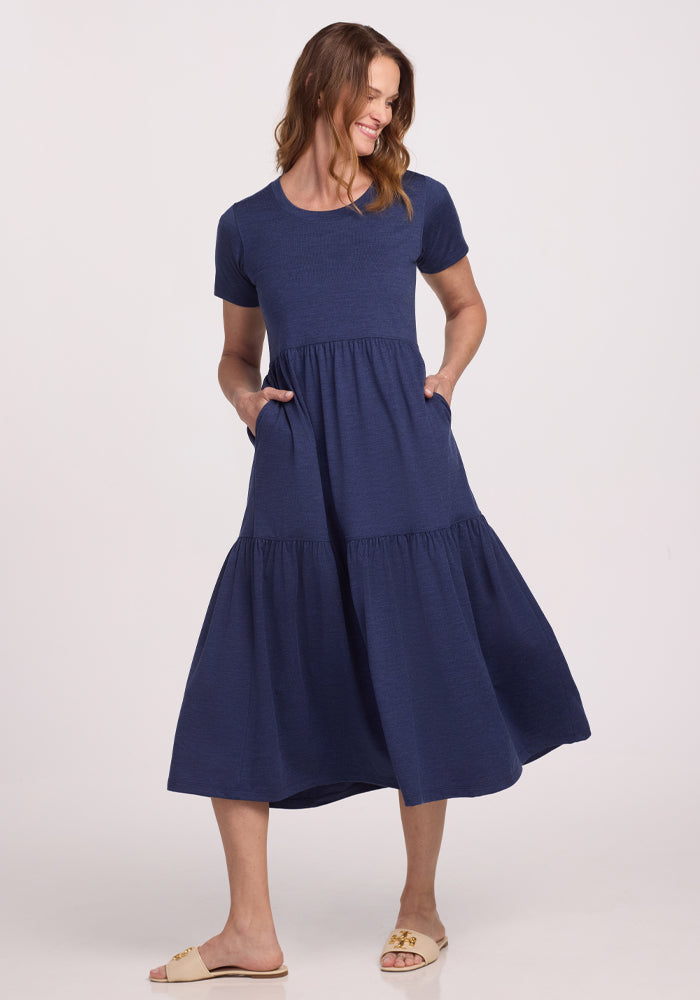 A person in a Woolx Lucia Dress - Twilight Storm stands against a white background. This short-sleeve, tiered design is ideal for summer. With hands in the pockets and beige sandals, they smile slightly while looking to the side.