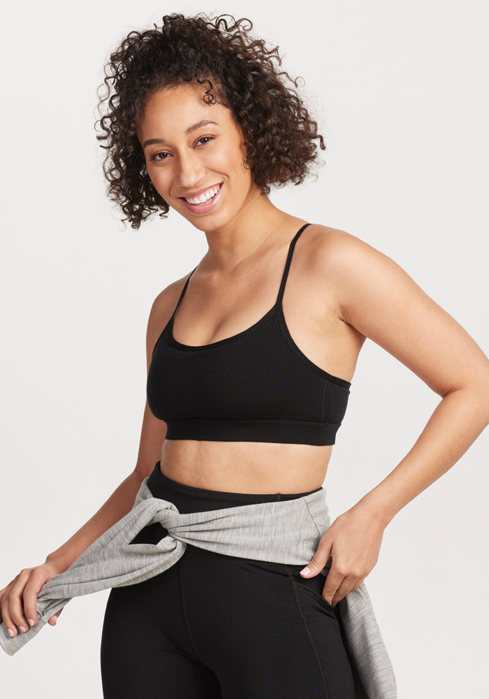 A person with curly hair smiles while wearing a black Luna Bralette by Woolx and moisture-wicking black leggings. They have a gray sweatshirt tied around their waist and stand against a plain background. | Tori is 5'7", wearing a size S
