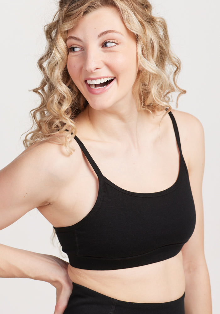 A person with curly blonde hair, smiling and looking to the side, is wearing the Woolx Luna Bralette in black. The plain, light-colored background perfectly highlights their cheerful expression. | Jordan is 5'6", wearing a size S