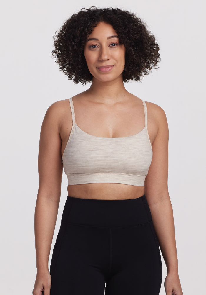 A woman with curly hair wears the Woolx Luna Bralette in Cream Heather and high-waisted black leggings, standing and smiling against a plain white background.