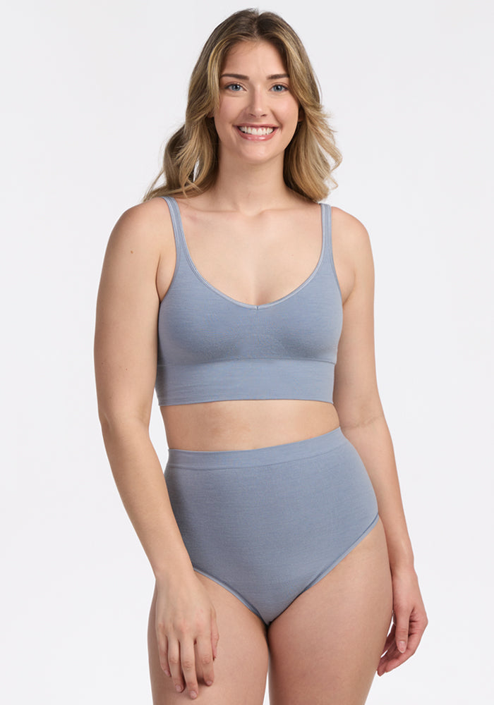 A woman with long, wavy blonde hair smiles at the camera while wearing the Woolx Mabel Full Coverage Underwear in Bluebell, showcasing the comfort and style of this seamless set against a plain white background.