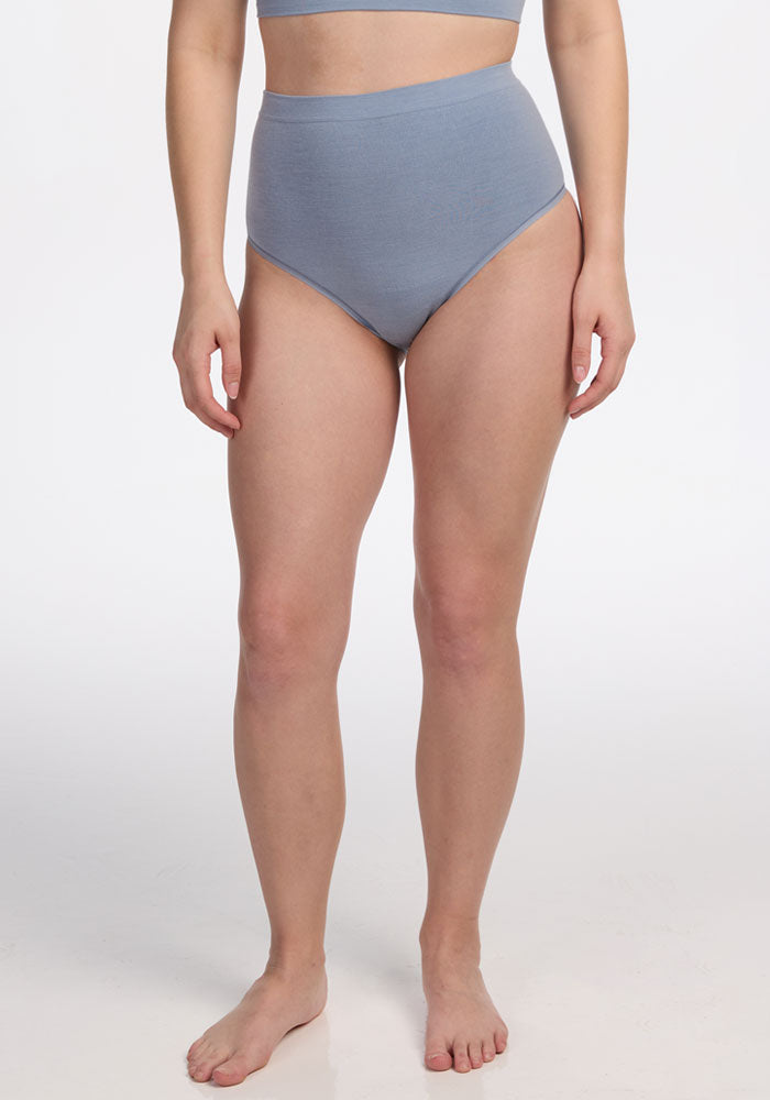 A woman stands barefoot on a white background, wearing Woolx’s Mabel Full Coverage Underwear in Bluebell with a matching crop top. Her arms are relaxed at her sides, and her face is not shown.