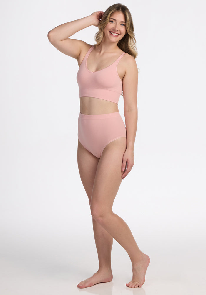 A woman with long blonde hair poses barefoot, smiling in Woolx’s Mabel Full Coverage Underwear in Rosewater and a matching sports bra, set against a plain white background. | Ariana is 5'11", wearing a size S