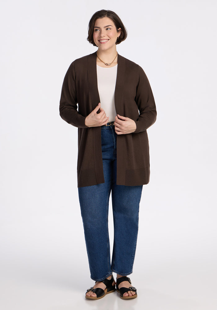 A woman with short brown hair, in a white top, blue jeans, black sandals, and the Woolx Macie Sweater - Bracken, smiles against a plain white background.