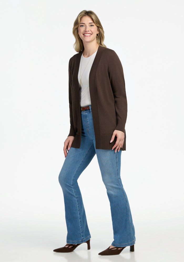 A person stands smiling against a white background, wearing the Woolx Macie Sweater in Bracken over a white top, blue flared jeans, brown heeled boots, and a necklace.