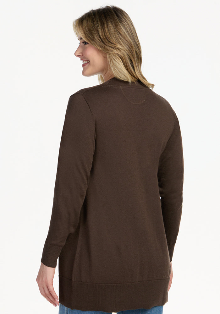 A woman with shoulder-length blonde hair, wearing the Woolx Macie Sweater in Bracken and blue jeans, stands smiling and looking to her left. She is shown from the back against a plain white background.